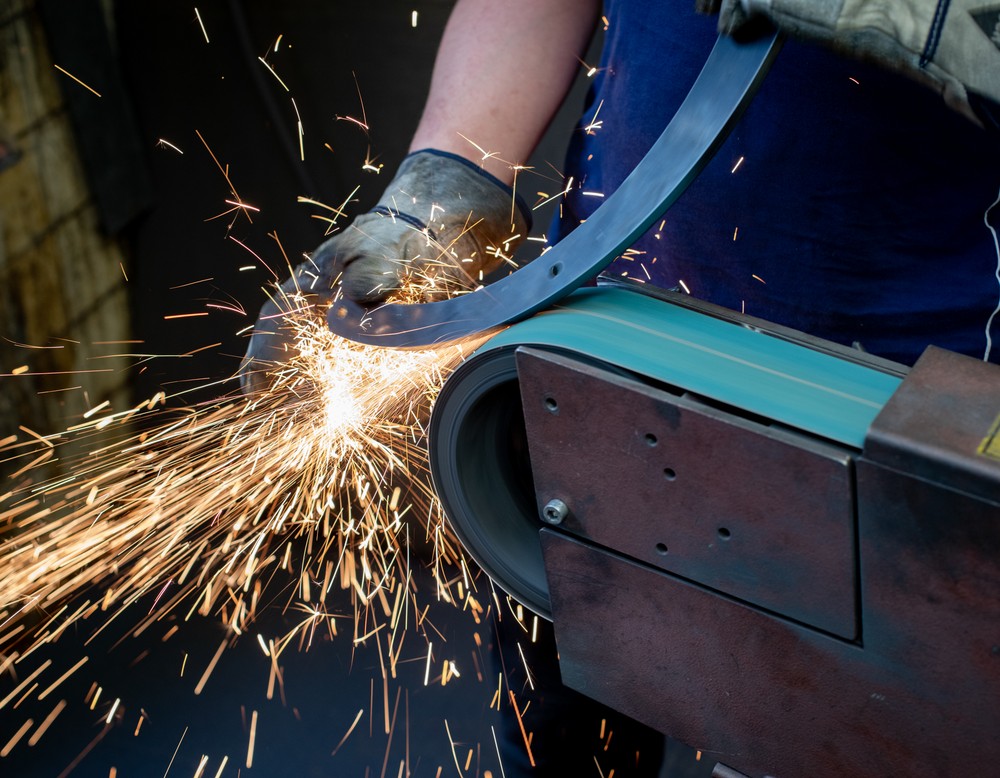 Zwei Hände im Arbeitshandschuhen schleifen Metall an einer Industriemaschine.